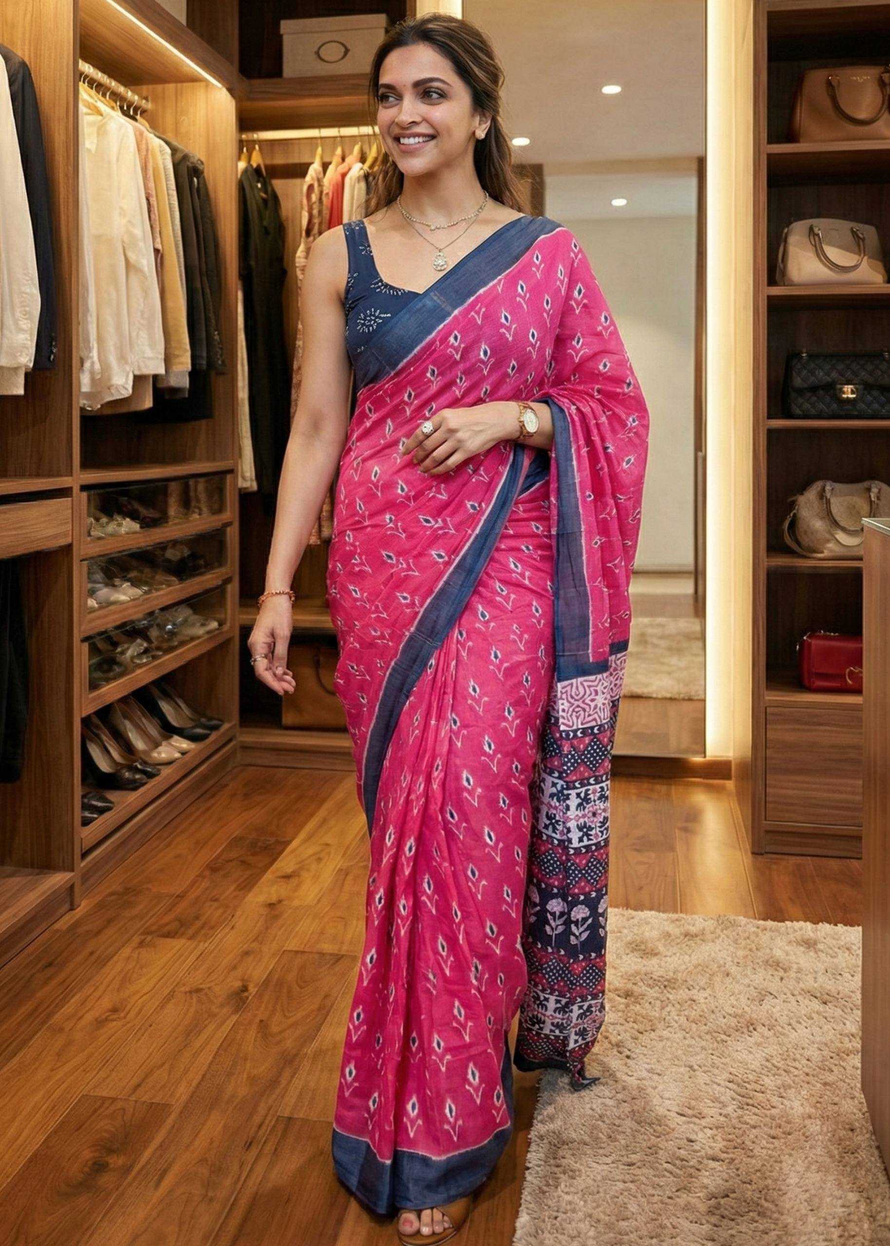 Woman in a pink saree with blue border standing in a well-lit room with wooden flooring.
