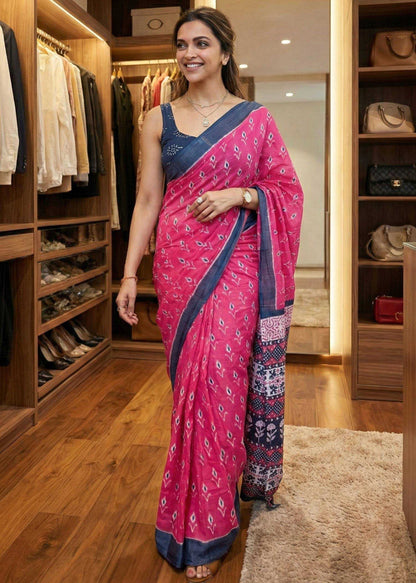 Woman in a pink saree with blue border standing in a well-lit room with wooden flooring.