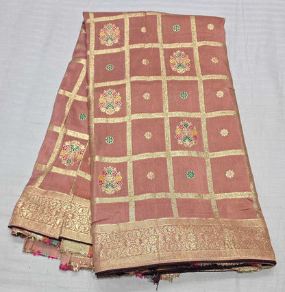 A folded terracotta pink Banarasi silk saree with gold zari borders, grid patterns, and colorful floral motifs, displayed on a light gray fabric background.