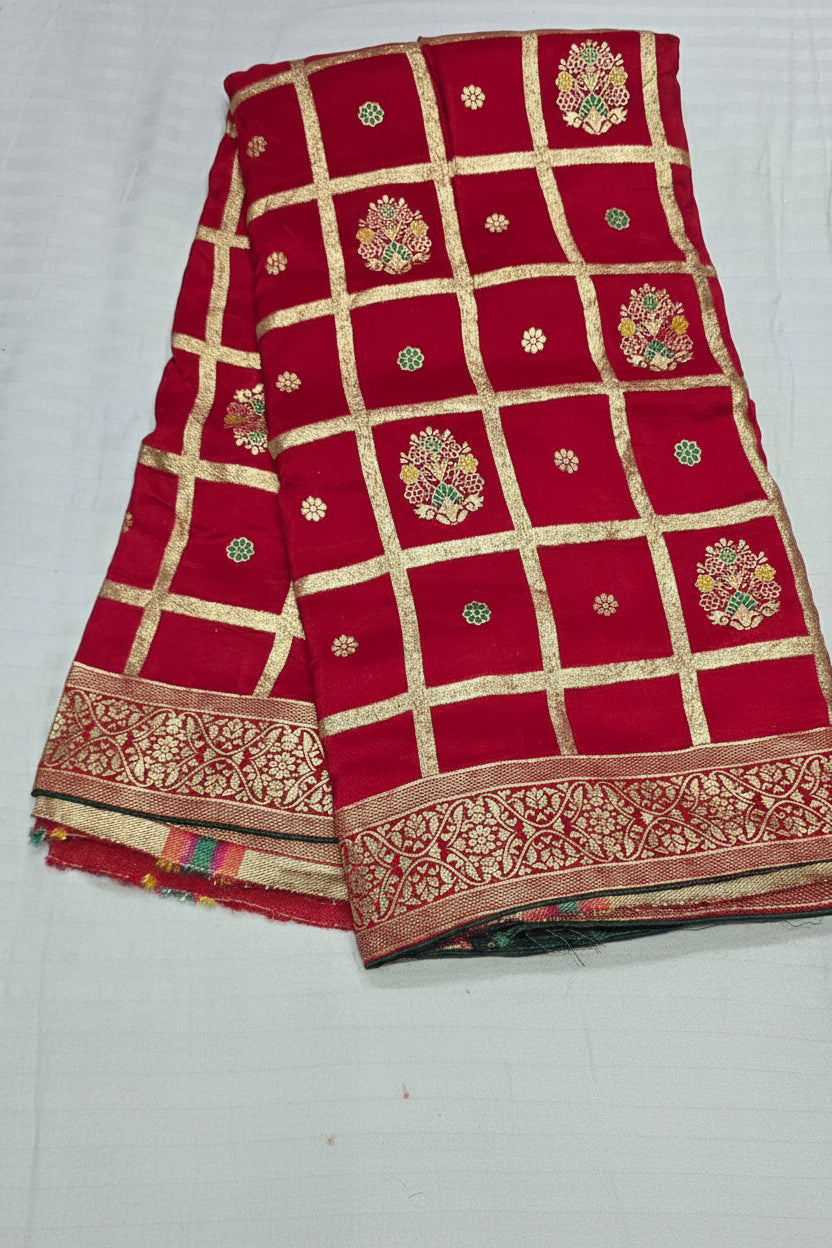 A folded red fabric, likely a saree or textile, featuring a gold grid pattern, ornate floral motifs, and a decorative gold border. A white text overlay in the center reads 'Red'.