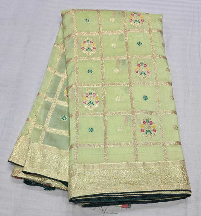 A folded light green silk fabric with gold zari borders and floral motifs, displayed on a light gray textured background.
