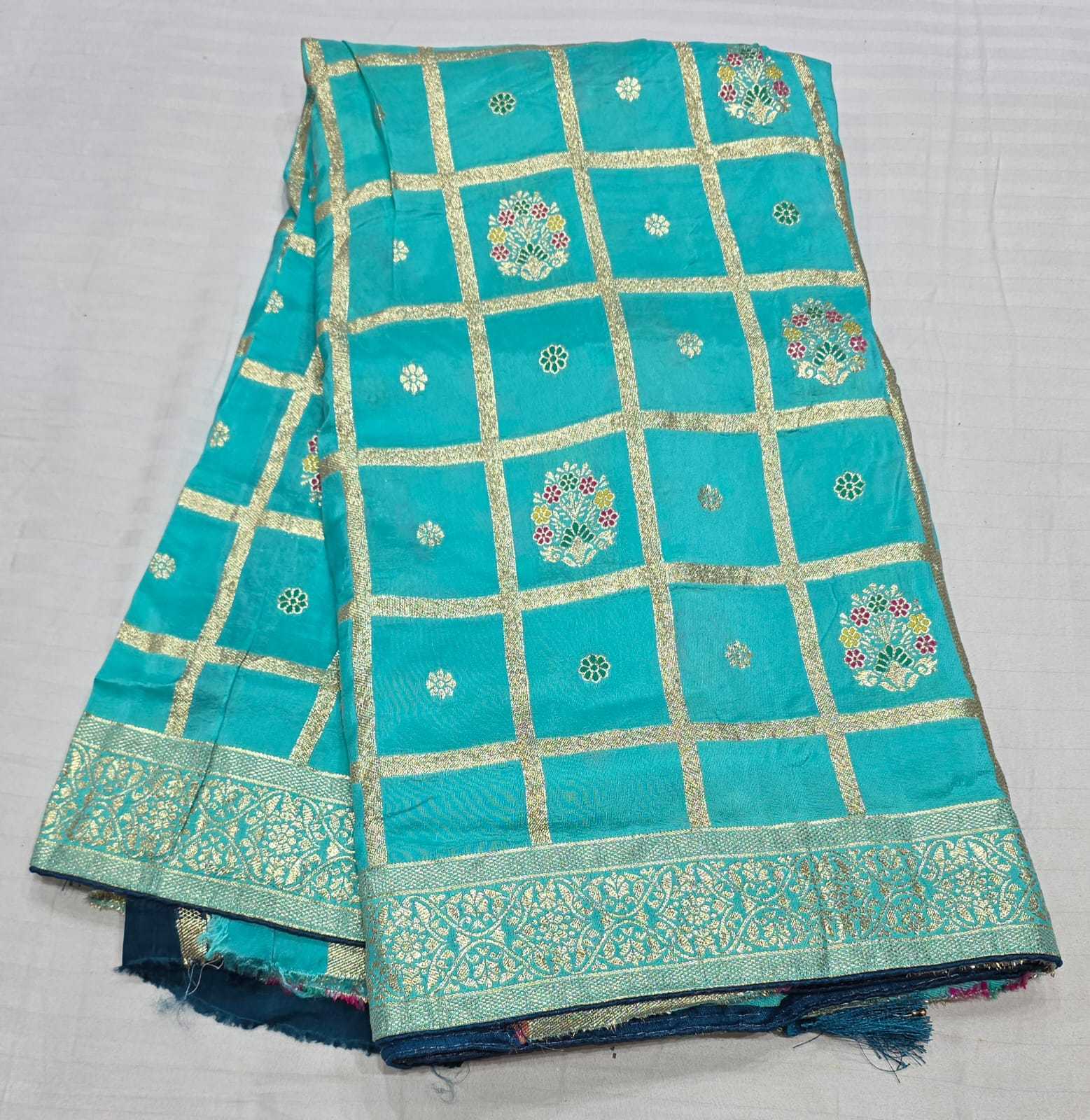 Turquoise blue Banarasi silk fabric with gold zari grid patterns, floral motifs, and a decorative border, folded on a white textured background.