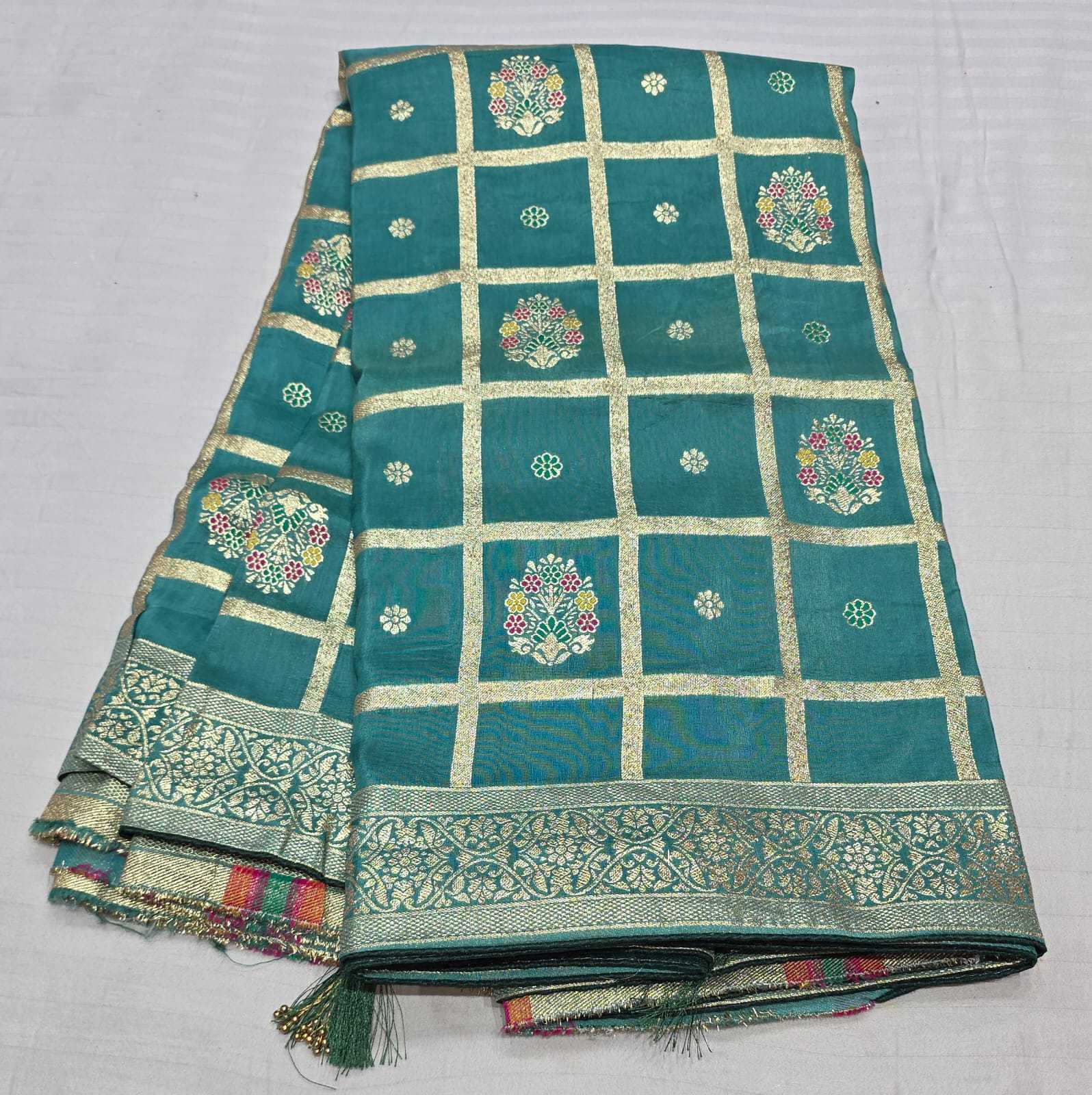 A folded teal green silk saree with intricate gold zari borders, grid patterns, and colorful floral motifs, displayed on a light gray fabric background.