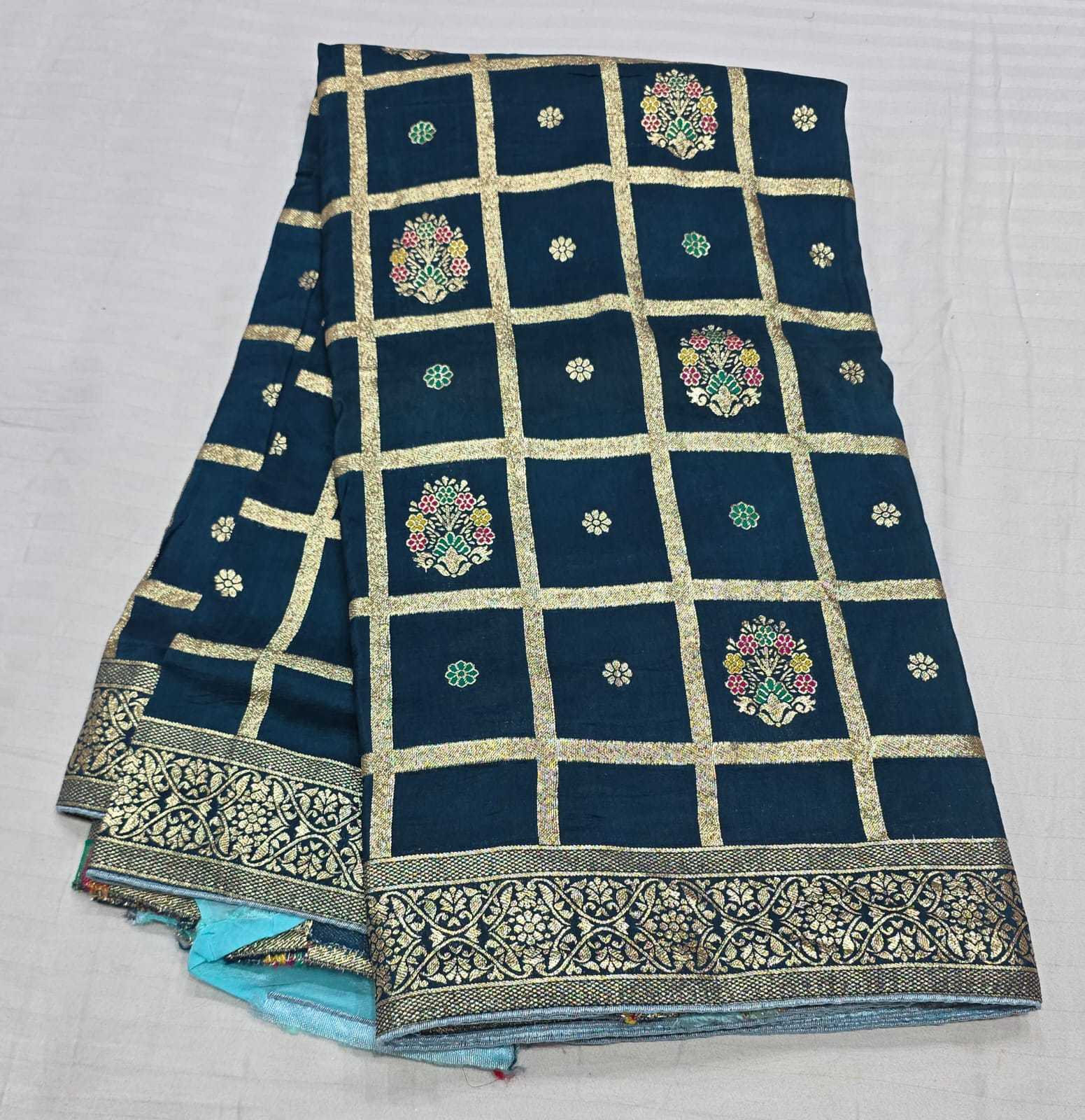 A folded dark teal blue fabric saree or textile piece with a gold grid pattern, colorful floral motifs, and an ornate gold border, displayed on a light gray fabric background.