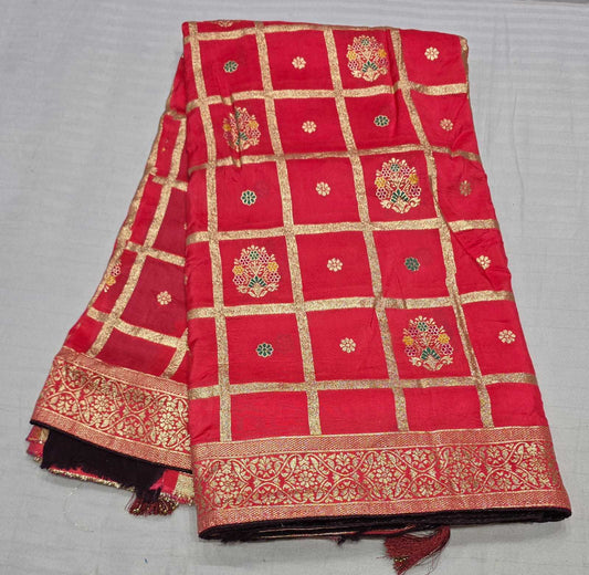 A folded red Banarasi silk saree fabric with gold zari grid patterns, floral motifs, and a decorative border, displayed on a light gray fabric background.