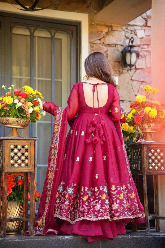Woman in a red embroidered dress standing outdoors with flowers and a stone wall in the background