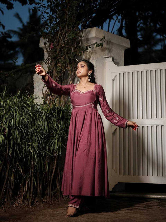 Woman in a maroon traditional outfit standing outdoors with a white gate and greenery in the background.